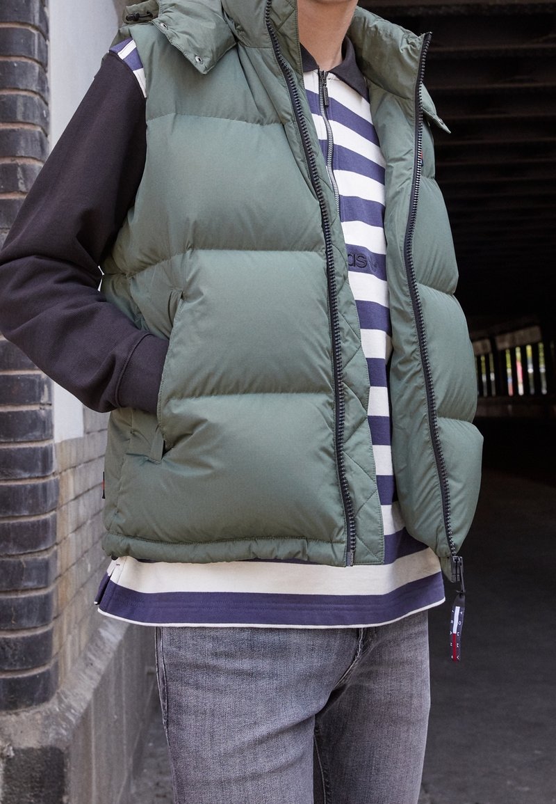Prošívaná vesta olivově zelené barvy se zipovým zapínáním a kapsami po stranách; nošena přes námořnicky modro-bílý pruhovaný svetr a šedé džíny.