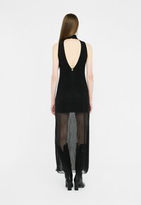 Abito nero senza maniche con profondo scollo a V sul retro, caratterizzato da un sovrapposto trasparente e delicate righe verticali. L'orlo presenta un design lungo e fluido, abbinato a stivali alla caviglia.