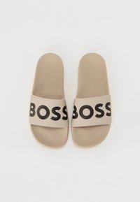 Beige Slide-Sandalen mit einer strukturierten Fußbett. Sie verfügen über einen breiten Riemen, der mit einem großen schwarzen "BOSS"-Logo verziert ist. Flaches und offenes Design.