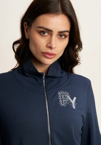 Giacca zip-up blu navy con colletto alto. Caratterizzata da un logo testurizzato "FY" adornato con strass sul lato sinistro. Design liscio e aderente.