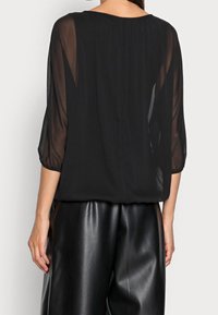 Czarna przezroczysta bluzka z rękawami 3/4, okrągłym dekoltem i luźnym krojem, zestawiona z błyszczącymi czarnymi spodniami w plisy.