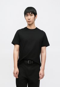 Čierne bavlnené tričko s klasickým okrúhlym výstrihom a krátkymi rukávmi, nosené s čiernymi nohavicami a jednoduchým opaskom, na bielom pozadí.