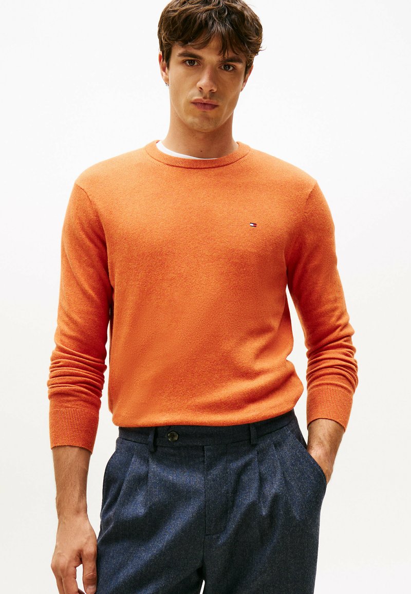 Orange Strickpullover mit Rundhalsausschnitt und langen Ärmeln. Mit einem kleinen Logo auf der Brust. Kombiniert mit dunklen strukturierten Hosen.