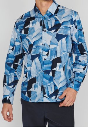 Camicia da uomo a maniche lunghe con bottoni e motivo astratto con pennellate blu, bianche e nere, indossata con pantaloni scuri.