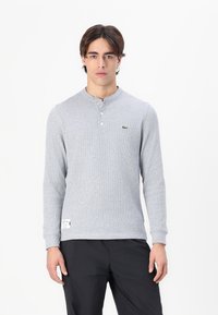 Sivi dolgi rokav henley majica z teksturirano tkanino, trije gumbi pri ovratniku in majhna zelena logotip na prsih.