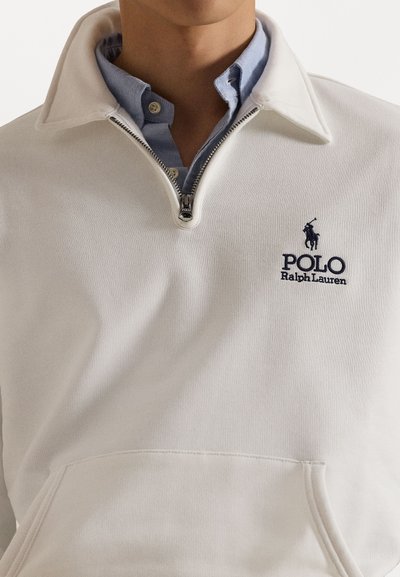 Valge fliisist veerandzipperiga pulover, millel on eestasku. All on sinine kaelusega särk ning tumesinise Polo Ralph Laureni logo.