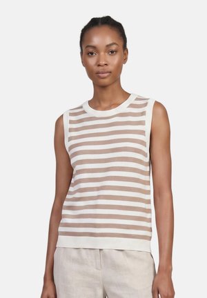 Donna che indossa un top beige e bianco a righe orizzontali senza maniche e pantaloni beige chiaro, in piedi davanti a uno sfondo bianco semplice.