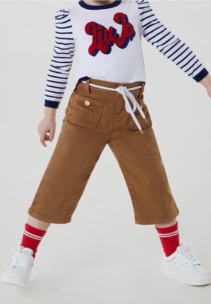 Pantalon court marron avec taille à cordon et poches latérales. Porté avec une chemise blanche ornée de lettres rouges scintillantes et de rayures bleu marine.