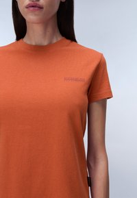 Oranges Baumwoll-T-Shirt mit Rundhalsausschnitt, kurzen Ärmeln und einem kleinen lila Logo auf der linken Brust. Weicher Stoff und lässige Passform.