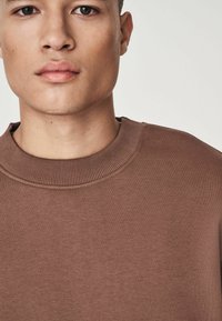 Sudadera marrón con cuello redondo acanalado, que presenta una textura suave y un detalle de costura mínimo, mostrando un diseño simple y estilizado.