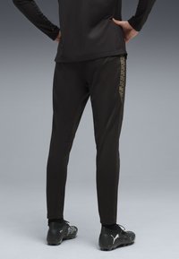 Svarta träningsbyxor med slim fit, prydda med ett mönstrat band längs sidan och svarta spikskor med dekorativa detaljer.