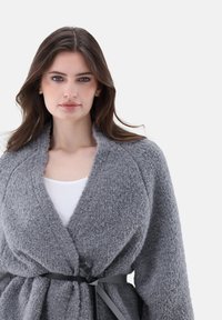 Grau gestrickte Strickjacke mit Schalkragen und einem gebundenen Gürtel in der Taille. Der Stoff scheint weich und gewebt zu sein, was Wärme und Komfort erhöht.