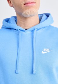 Světlě modrá mikina Nike vyrobená z měkké látky, s přední kapsou, stahovacími šňůrkami a malým bílým logem na levé straně hrudi.