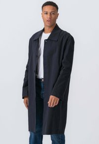 Mand iført en sort lang frakke over en hvid skjorte og blå jeans, som står imod en ensfarvet lys grå baggrund.