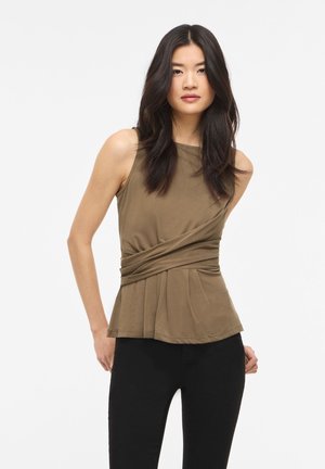 Femme aux longs cheveux bruns portant un haut olive sans manches avec un détail froncé à la taille et un pantalon noir, debout devant un fond blanc uni.