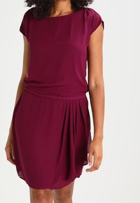 Bordeaux Kleid mit kurzen Ärmeln, plissiertem Rock und tailliertem Schnitt. Hergestellt aus leichtem Stoff mit glatter Textur und subtiler Brillanz.