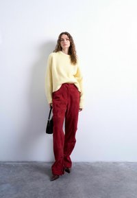 Femme debout contre un mur blanc, portant un pull jaune, un pantalon large en velours rouge, des chaussures pointues marron, et tenant un sac à main noir.