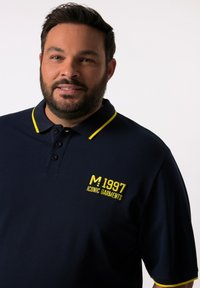 Marinblå polo-tröja med gul krage och detaljer, med en broderad logotyp "M 1997 ICONIC GARMENTS" på vänster bröst. Slät textur.