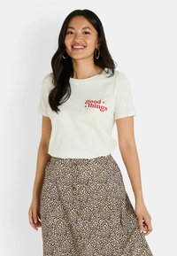 T-shirt crème à manches courtes avec un imprimé graphique rouge "good things", associé à une jupe beige à motifs léopard avec des détails en boutons.