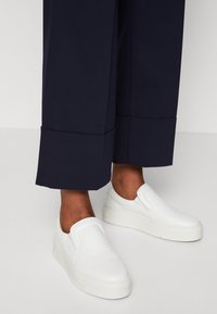 Białe sneakersy slip-on o eleganckim designie, z teksturowaną skórzaną cholewką i grubą białą podeszwą, zestawione z granatowymi spodniami typu cropped.