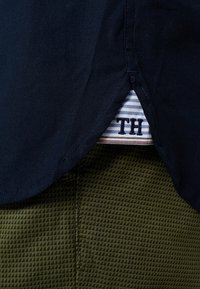 Camicia blu navy con orlo curvo, che mostra un'etichetta a righe con "TH". Tessuto verde testurizzato dei pantaloni visibile sotto l'orlo della camicia.