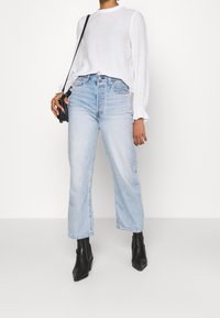 Blouse blanche légère à manches longues avec des poignets à volants, associée à un jean taille haute bleu clair à jambes larges, et des bottines noires à bout pointu.