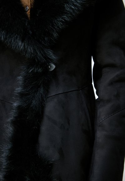 Gros plan d’un manteau noir avec une bordure en fourrure douce et duveteuse le long du devant et un bouton visible près du col.