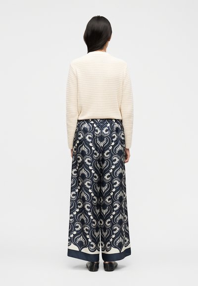 Γυναίκα με μακριά σκούρα μαλλιά φοράει κρεμ πουλόβερ με υφή και φαρδιά μπλε παντελόνια με λευκό λουλουδάτο σχέδιο, στέκεται μπροστά σε λευκό φόντο.