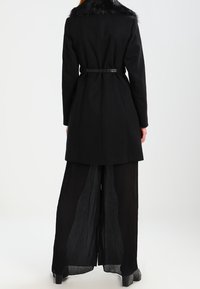 Svart yllejacka med krage av fuskpäls, midjebälte som dras åt och långa, plisserade svarta byxor. Slät textur med skräddarsydd passform.