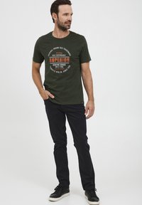 Mørkegrønn bomulls t-skjorte med rund hals og oransje tekstdesign, sammen med svarte bukser og svarte joggesko. Korte ermer, vanlig passform.
