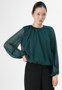 Woman wearing dark green sheer zigzag patterned blouse with long sleeves and black pants, standing with one hand on hip.