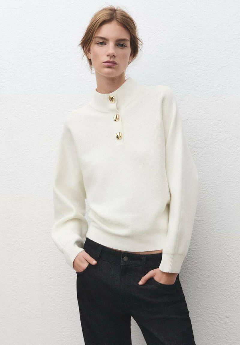 Jeune femme portant un pull blanc avec trois boutons dorés au col et un pantalon noir, debout devant un mur blanc texturé.