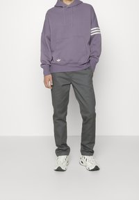 Sweat à capuche violet avec une poche avant, orné de bandes blanches sur les manches et un petit logo. Associé à un chino gris et des baskets blanches.