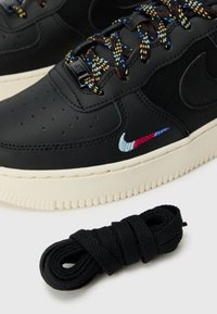 Baskets noires avec des lacets multicolores, semelle blanche, logo Nike brodé en rouge, bleu et blanc, et un lacet noir enroulé sur une surface blanche.