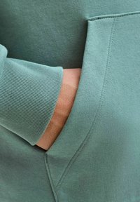 Mintgrünes Sweatshirt mit strukturiertem Finish, ausgestaltet mit einer Kängurutasche und gerippten Bündchen. Nahaufnahme, die die Nähdetails hervorhebt.
