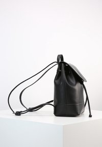Sac à dos en cuir noir avec un fond rond, fermeture à cordon et bretelles ajustables. Texture lisse et design minimaliste.