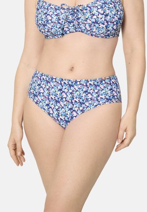 Mujer con bikini azul floral de braguita de talle alto y top con lazo, posando frente a un fondo blanco liso.