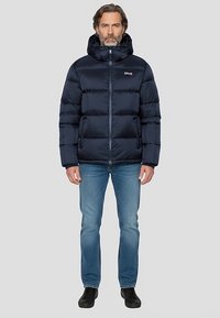 Veste matelassée bleu marine avec fermeture éclair et capuche, présentant un design matelassé et une légère brillance. Associée à un jean bleu et des chaussures noires.