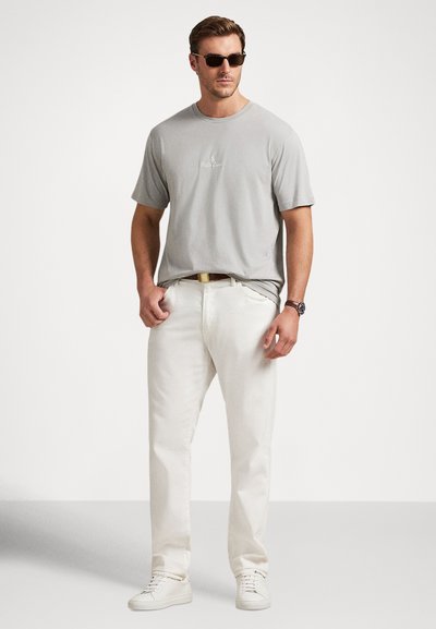 Mann iført lysegrå Ralph Lauren t-skjorte, hvite bukser, hvite joggesko, brunt belte, klokke og svarte solbriller, stående mot ensfarget bakgrunn.