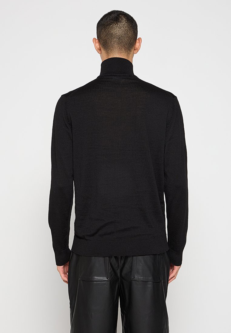 Homme avec des cheveux courts portant un pull à col roulé noir à manches longues et un pantalon en cuir noir, se tenant de dos devant un fond blanc.