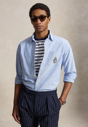 Fiatal férfi napszemüvegben, világoskék inggel, amin mackós folt található, csíkos sötétkék nadrágban és karórával, áll, egyik kezével a zsebében.