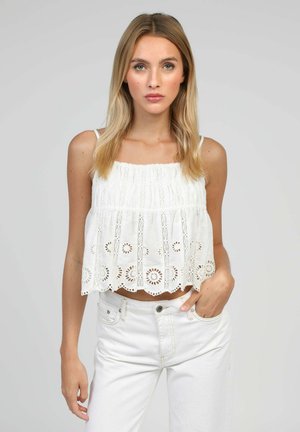 Jeune femme aux cheveux blonds raides porte un crop top blanc sans manches à œillets et un jean blanc taille haute, posant avec une main dans la poche.