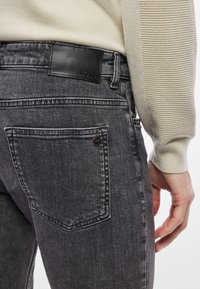 Σκούρο γκρι τζιν με απαλό υφή, που διαθέτει μαύρο δερμάτινο λογότυπο και δύο πίσω τσέπες με διακριτικές ραφές.