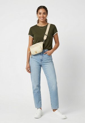 Frau lächelt, trägt ein olivgrünes T-Shirt, hellblaue Jeans, weiße Sneakers und eine beige Umhängetasche mit verstellbarem Riemen.