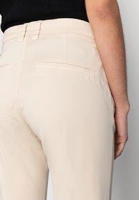 Calças bege com uma textura lisa, apresentando uma cintura ajustada e dois bolsos traseiros, mostradas de um ângulo lateral.