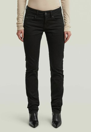 Persoon die strakke zwarte broek, beige top met lange mouwen en zwarte puntige enkellaarsjes draagt, staand tegen een effen lichte achtergrond.