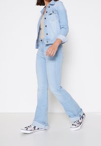 Chaqueta de mezclilla azul clara con bolsillos delanteros y botones, combinada con jeans azul claros y zapatillas con un patrón blanco y negro.