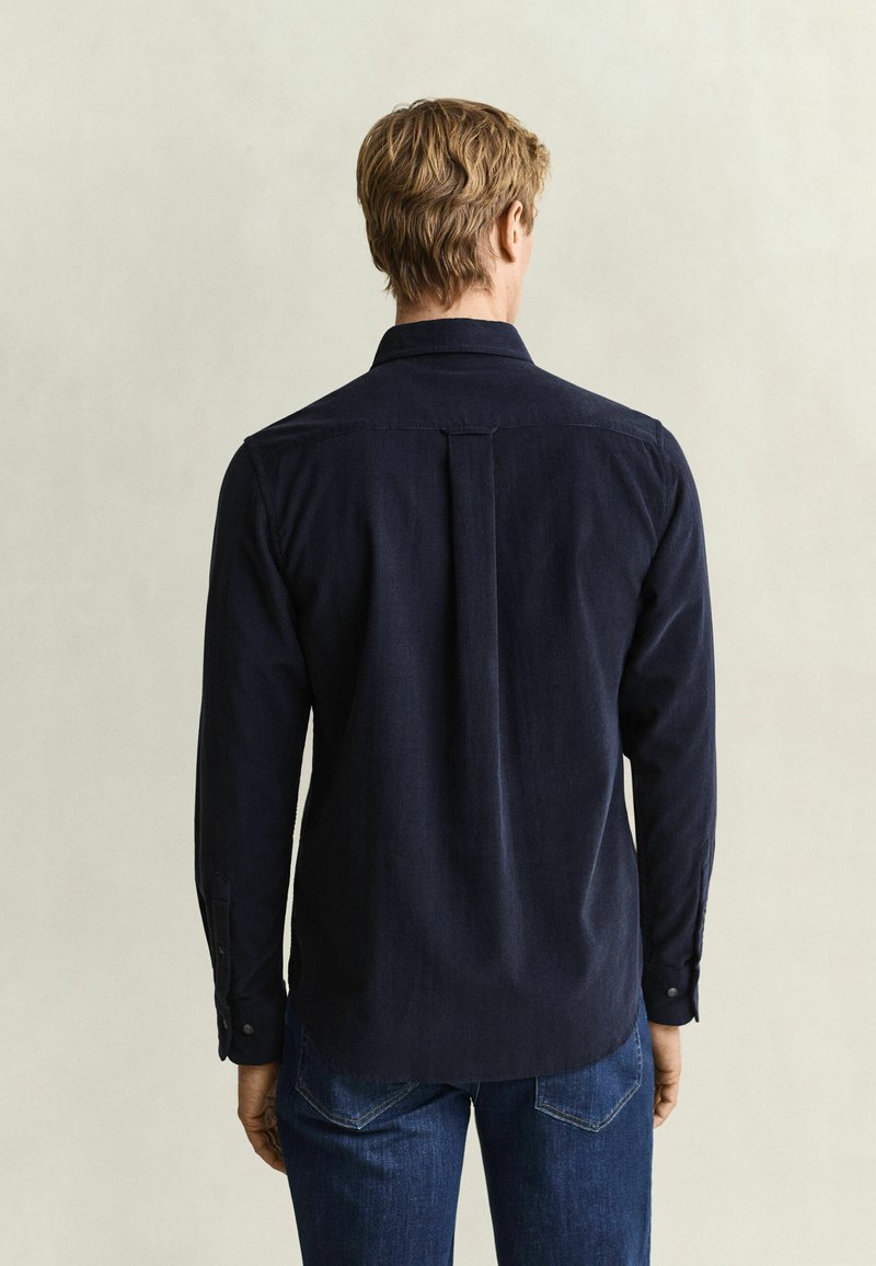Man with light brown hair wearing a dark long-sleeve shirt and blue jeans, standing facing a plain light background.