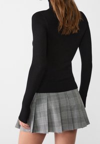 Femme portant un haut noir ajusté à manches longues et une mini-jupe plissée à carreaux gris, vue de dos sur un fond uni.