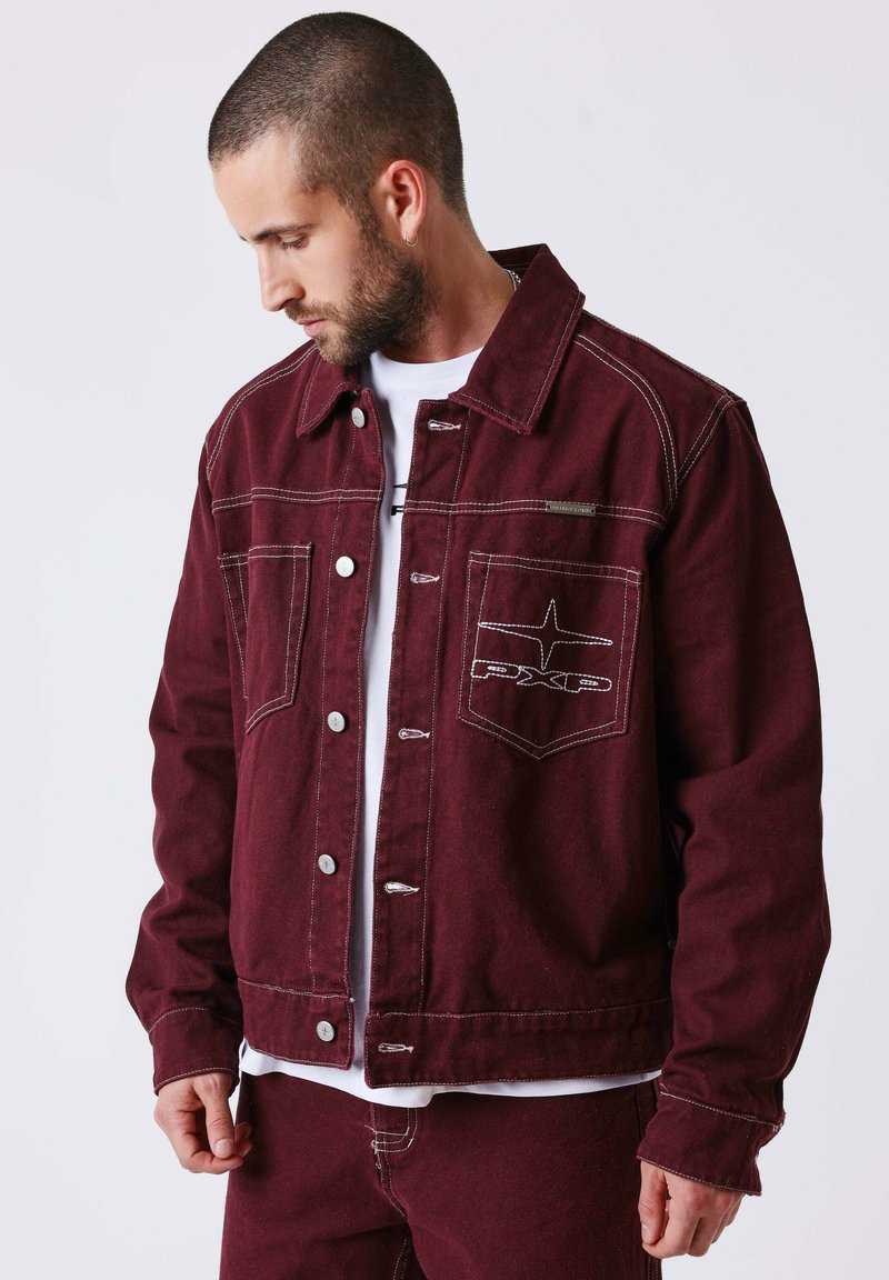 Homme à la barbe taillée portant une veste boutonnée bordeaux et un pantalon assorti, regardant vers le bas sur un fond uni.